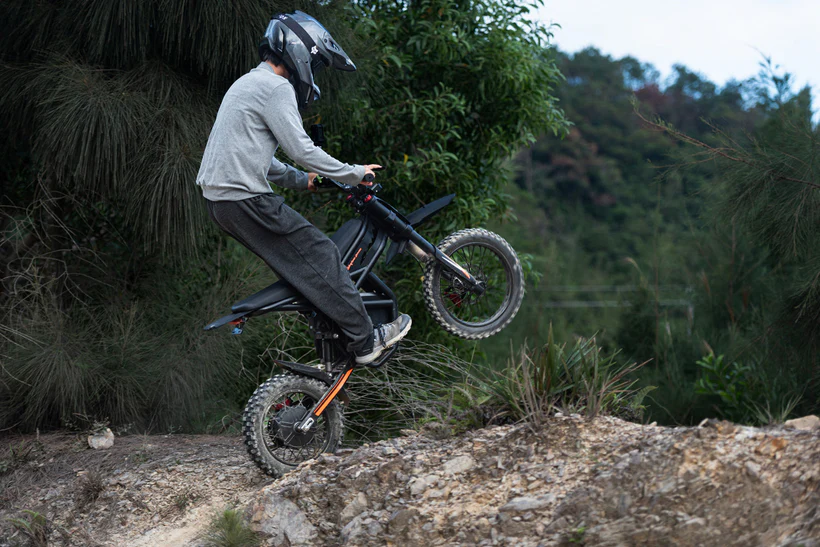 GT54 PRO elektrinis dviratis 1000W bekelėje – vairuotojas su šalmu kyla į stačią įkalnę ir atlieka „wheelie“, demonstruojant stiprų sukibimą, galingą variklį ir puikų pravažumą miško trasoje