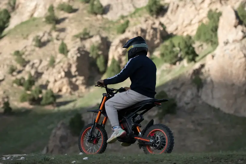 Vairuotojas su šalmu važiuoja GT54 elektriniu dviračiu kalnų ir žalių slėnių bekelėje, juodai oranžinis 1000W e-bike stabiliai įveikia nelygų reljefą gamtoje.